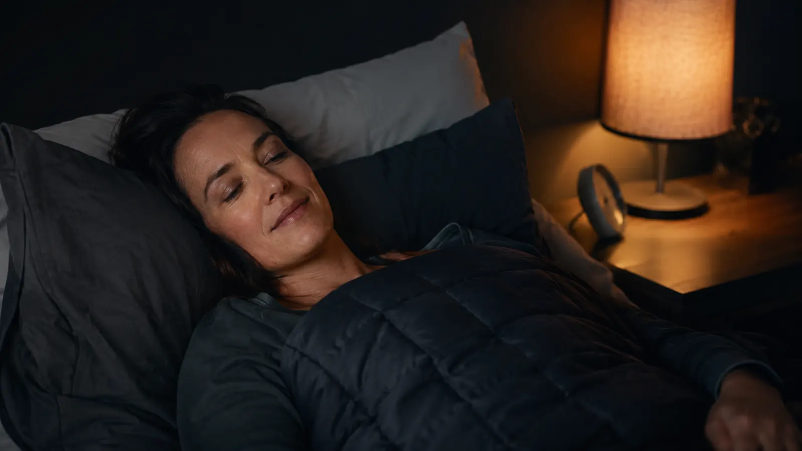 Adult woman lying in bed at night cocooned under a heavy weighted blanket, soft warm bedside lamp light, relaxed expression