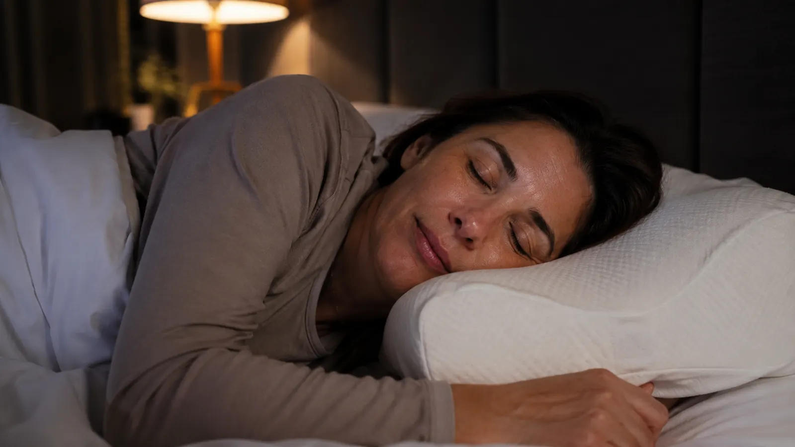 Woman sitting on edge of bed in morning light, holding the back of her neck with a tired expression