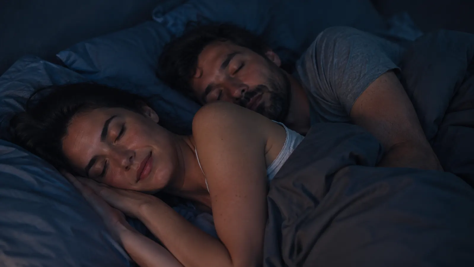 Person sleeping peacefully in a dark bedroom, white noise machine glowing softly on the nightstand