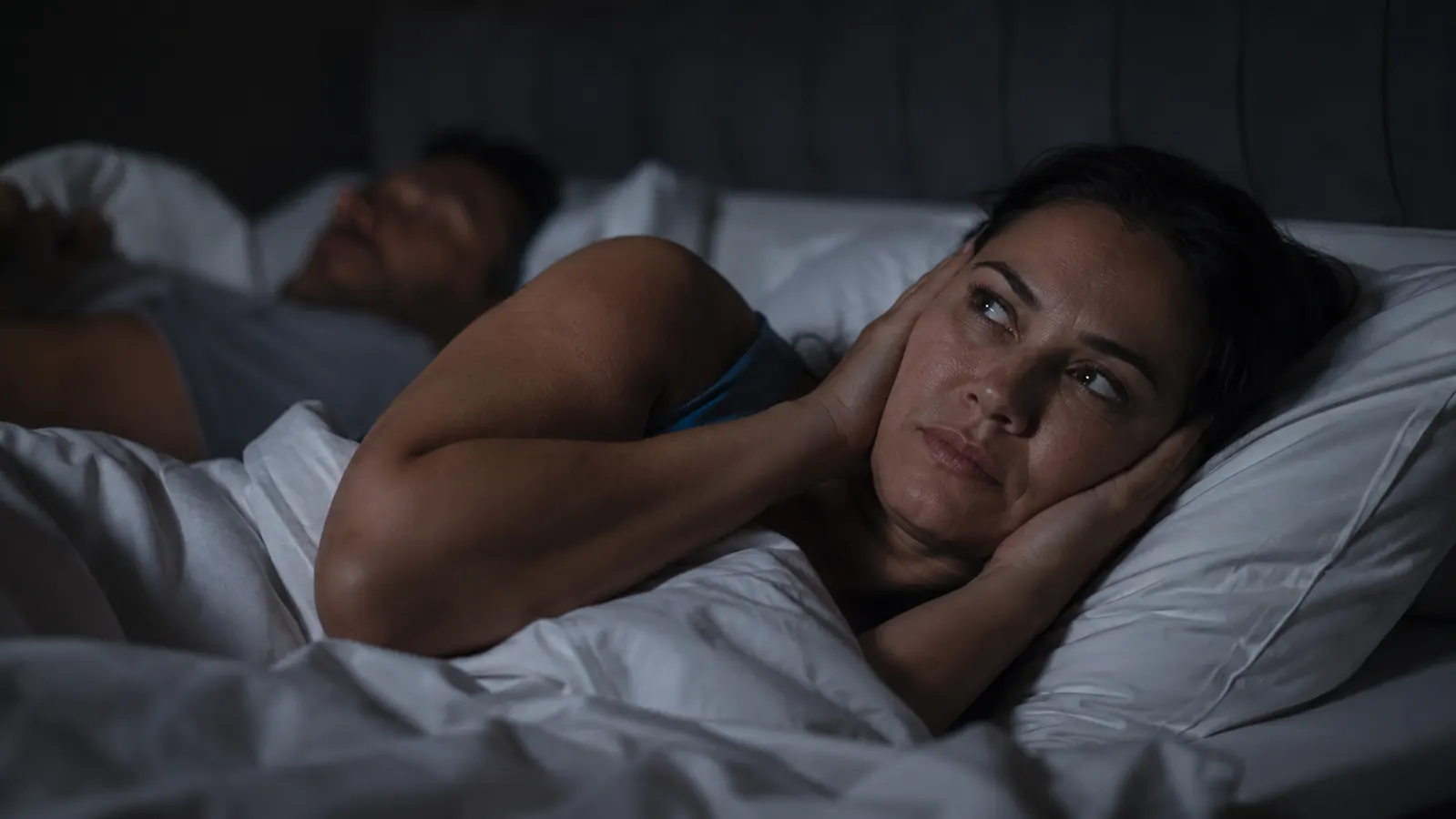 Person lying awake in a dark bedroom, partner asleep beside them, soft blue light through window blinds