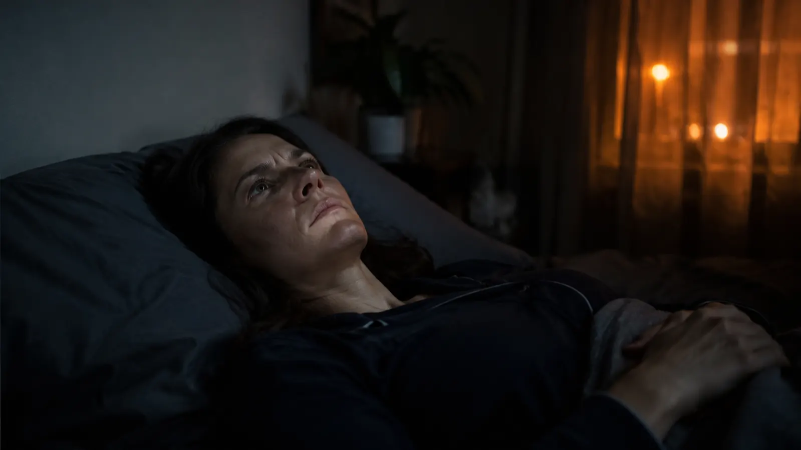 Woman lying awake in a dark bedroom, streetlight visible through curtains, partner asleep beside her